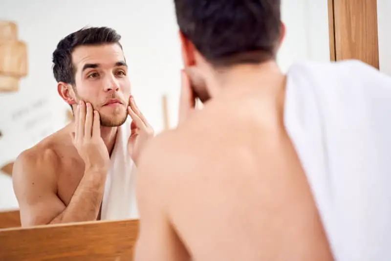 homem olhando para o espelho com a mao na barba Como ter barba cheia guia completo